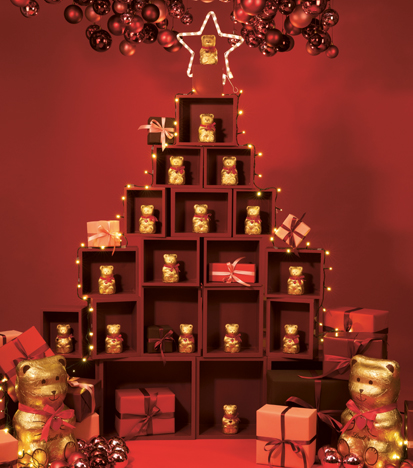 Lindt Teddies surrounded by Christmas decorations in front of a red background (Photo)