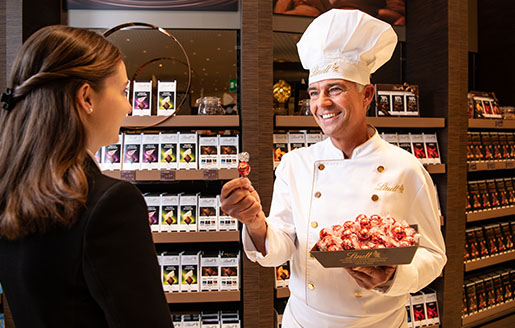 Maître chocolatier Urs Liechti offering a red LINDOR truffle to a female customer in a shop (Photo)