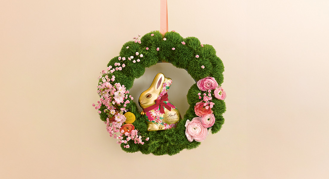 A Lindt Goldhase in a wreath with pink flowers. (Photo)