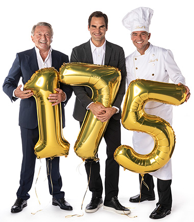 Ernst Tanner, Roger Federer und ein Maitre Chocolatier mit einem Glas Wein in der Hand. Vor ihnen steht ein Luftballone in der Form der Zahlen 175 (Foto)