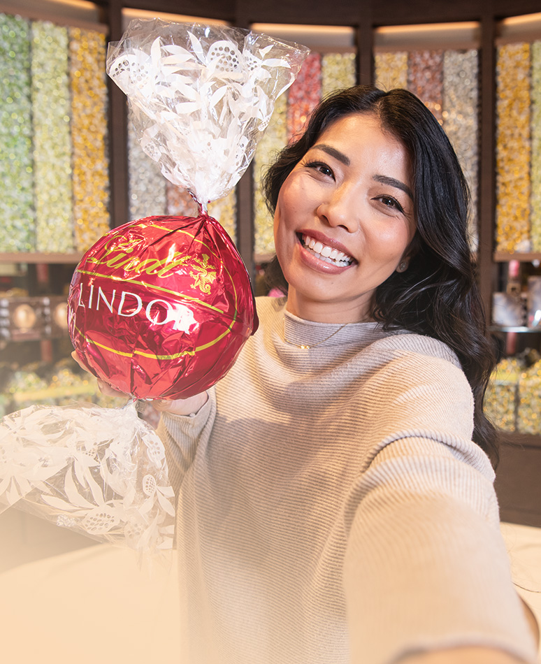 Eine braunhaarige, lächelnde Frau in einem Lindt-Shop mit einer großen roten LINDOR-Kugel in den Händen; im Hintergrund sind Behälter mit verschiedenen LINDOR-Kugeln (Foto)