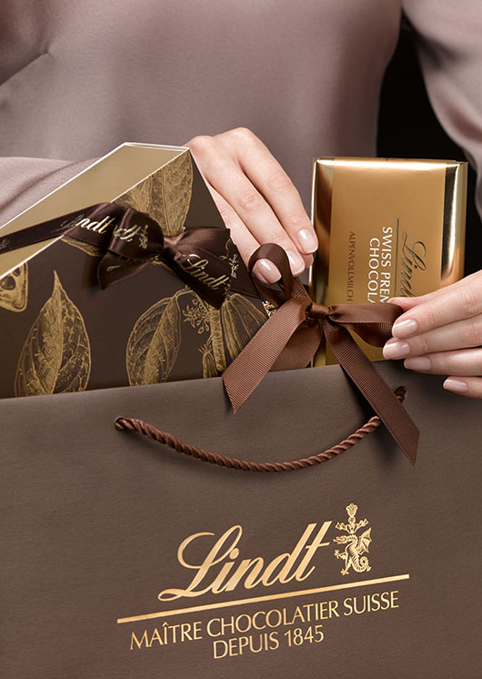 Eine braune Lindt-Geschenktasche mit diversen Lindt-Produkten; die eine weibliche Hand richtet eine braune Schleife, die andere die Schokoladentafel (Foto)