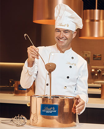 Maître chocolatier Urs Liechti hält einen Schöpflöffel in der Hand und schöpft Schokolade aus einem goldenen Topf (Foto)